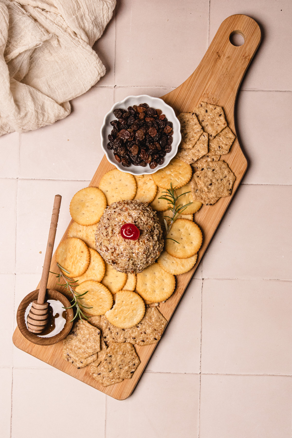 A wooden board with crackers, a pineapple cheese ball recipe topped with a cherry, honey dipper, rosemary, and a bowl of raisins.