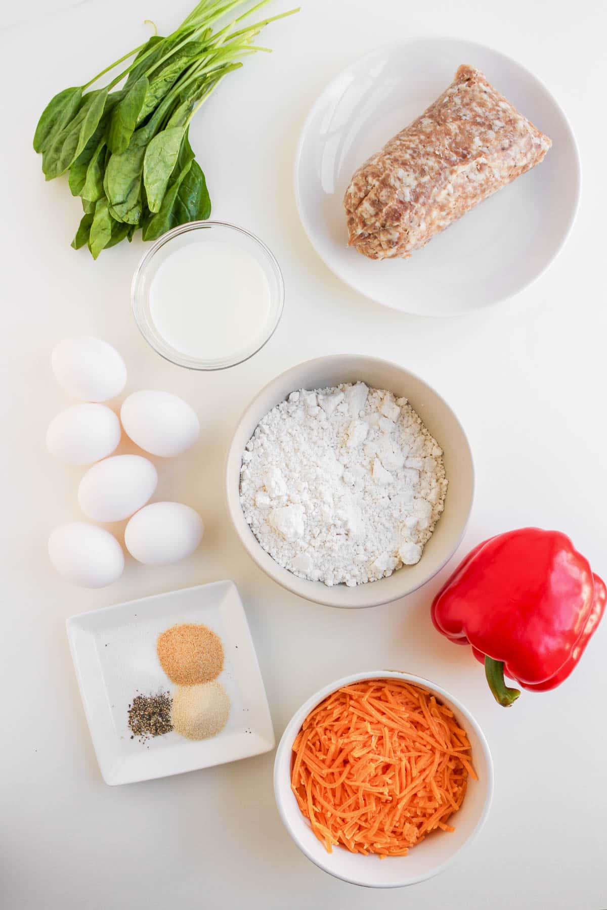 Ingredients on a white surface: eggs, spinach, milk, ground meat for sausage, flour, shredded carrot, red bell pepper, and spices—perfect for making savory Cheese Breakfast Muffins.