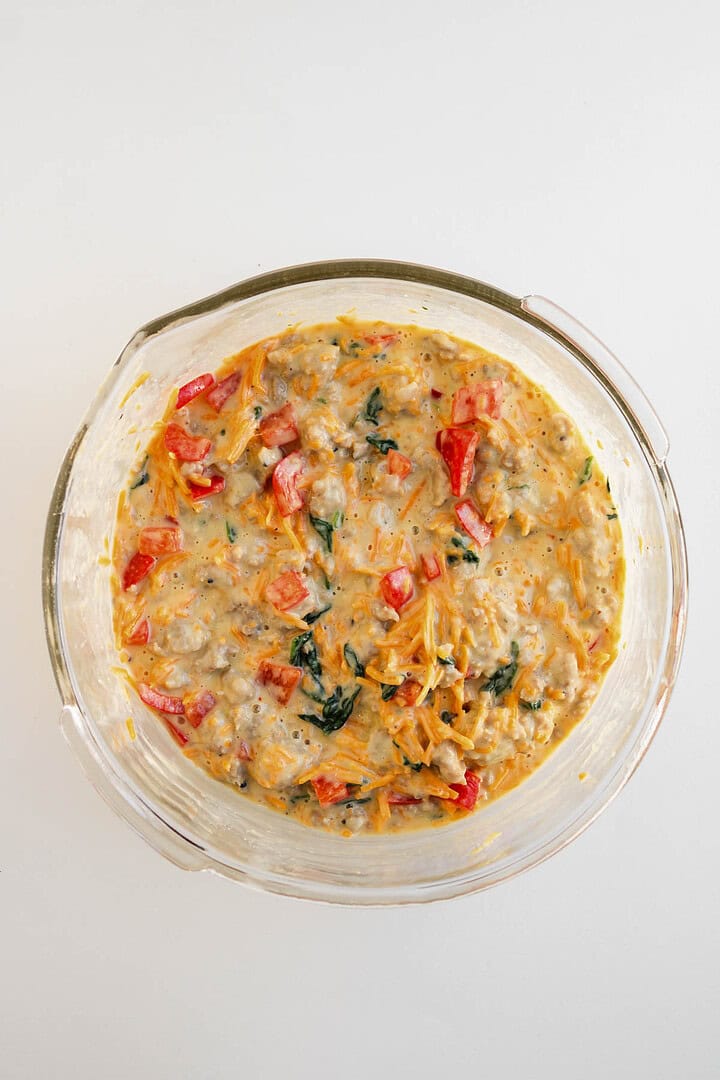 A glass bowl filled with a colorful, mixed vegetable and cheese batter—perfect for making savory egg and cheese muffins—sits on a white surface.