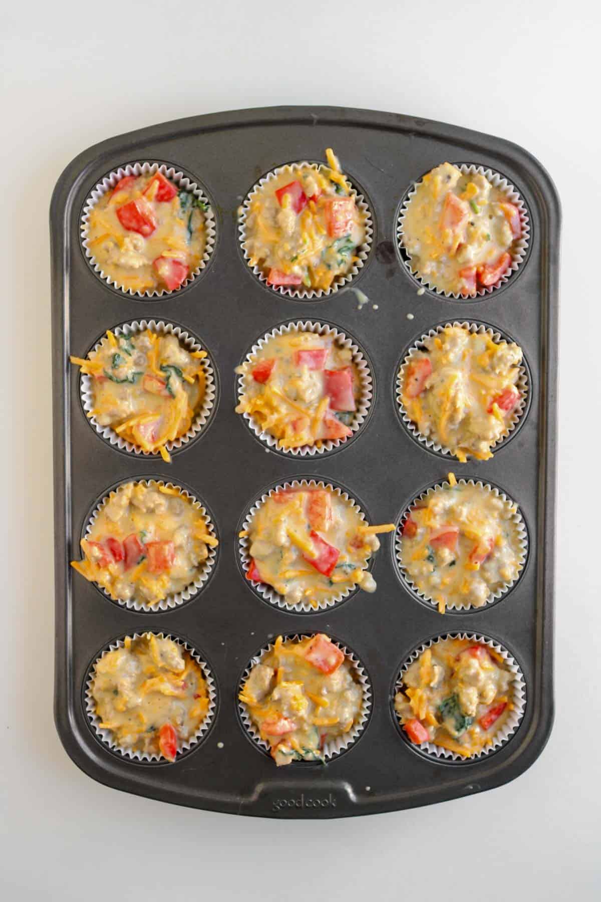 A muffin tin filled with unbaked breakfast muffins batter, featuring visible chopped vegetables, cheese, and bits of sausage ready to become delicious sausage breakfast muffins.