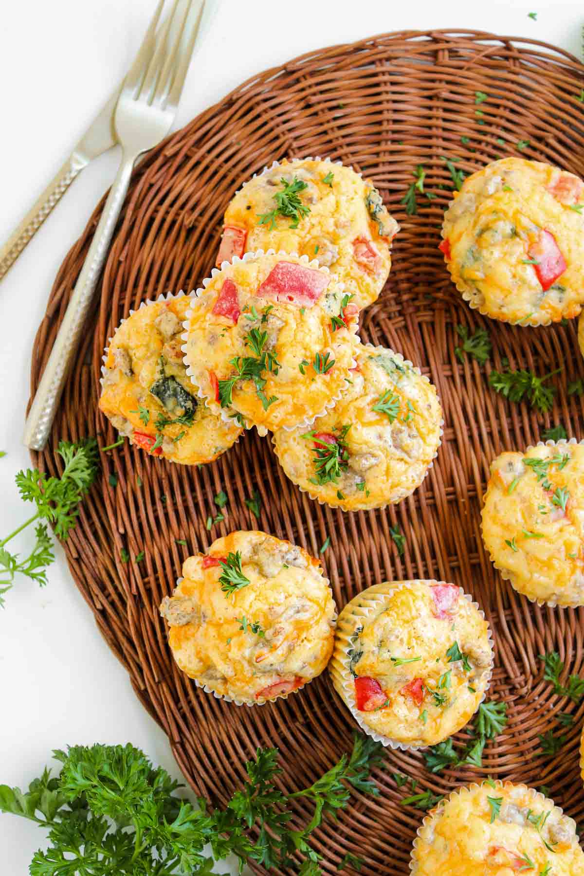 Egg and cheese muffins with vegetables and herbs on a wicker tray, garnished with parsley, two forks beside them—these savory breakfast muffins are perfect for a hearty start to your day.