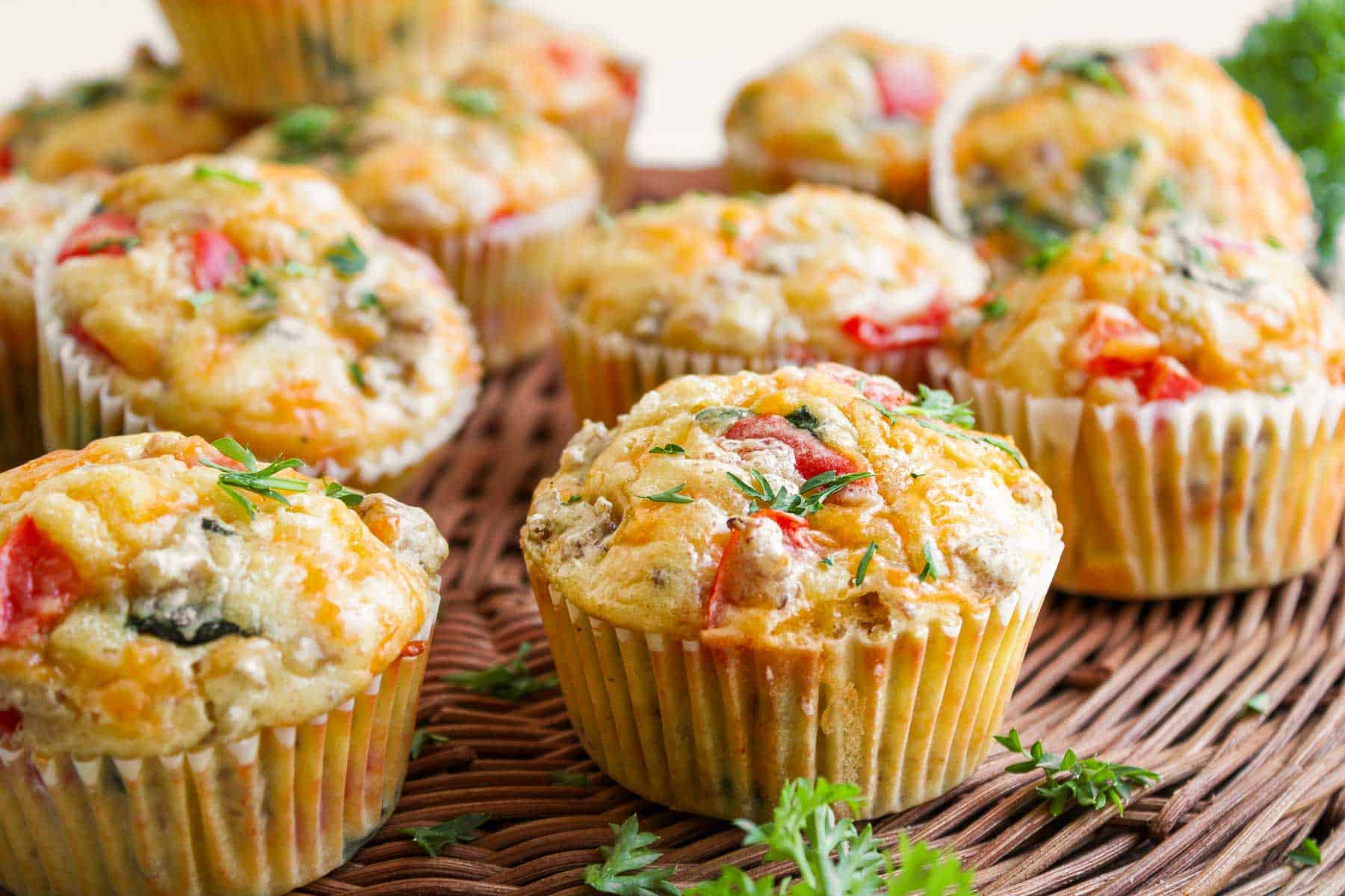 Savory breakfast muffins with vegetables, herbs, egg and cheese in paper liners, arranged on a woven tray.