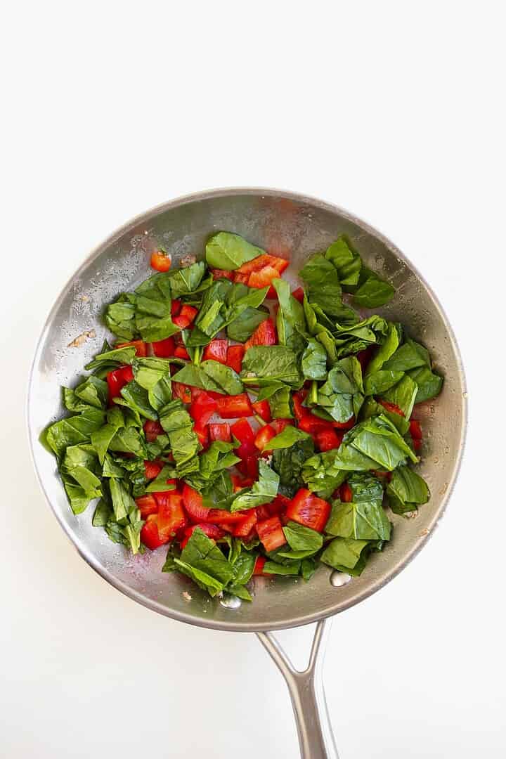 Chopped spinach, red bell pepper, and sausage sizzle in a metal frying pan—perfect ingredients for egg breakfast muffins—set on a white background.