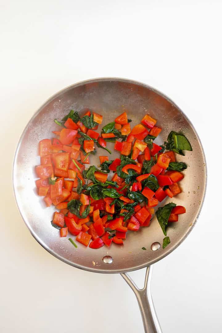 Diced red bell peppers and spinach sautéing in a stainless steel skillet on a white background, perfect for adding to savory Breakfast Muffins or pairing with classic Egg and Cheese.
