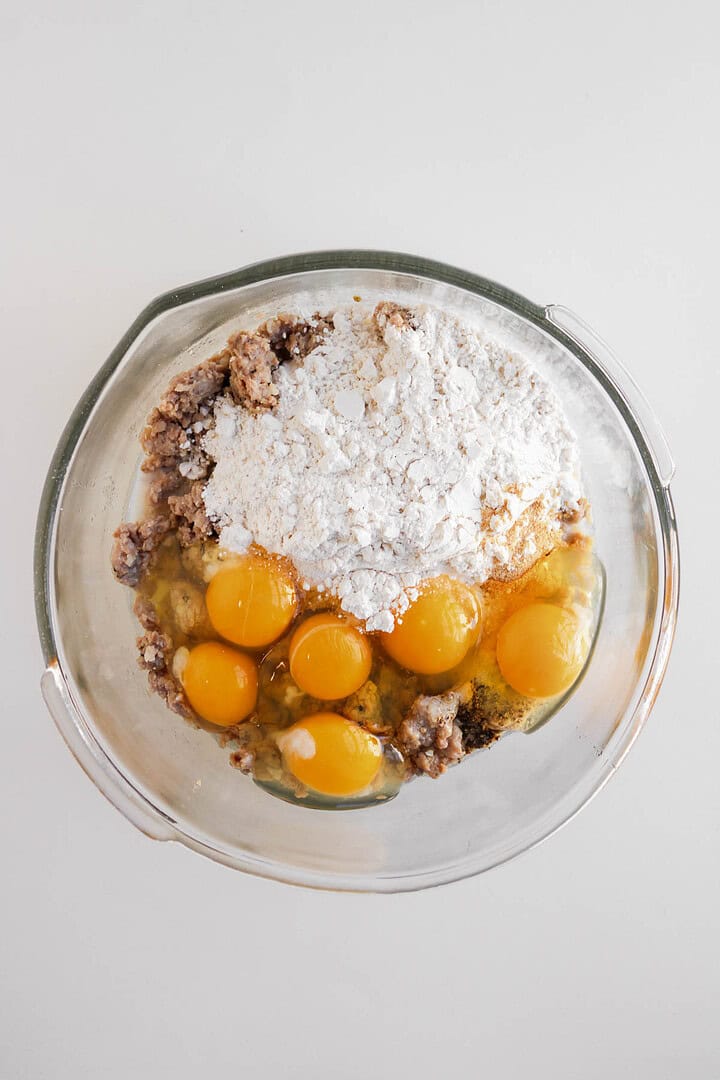 Glass bowl with cracked eggs, flour, and a brown mixture, ready to be mixed on a white surface—perfect for making savory egg and cheese muffins or hearty sausage breakfast muffins.