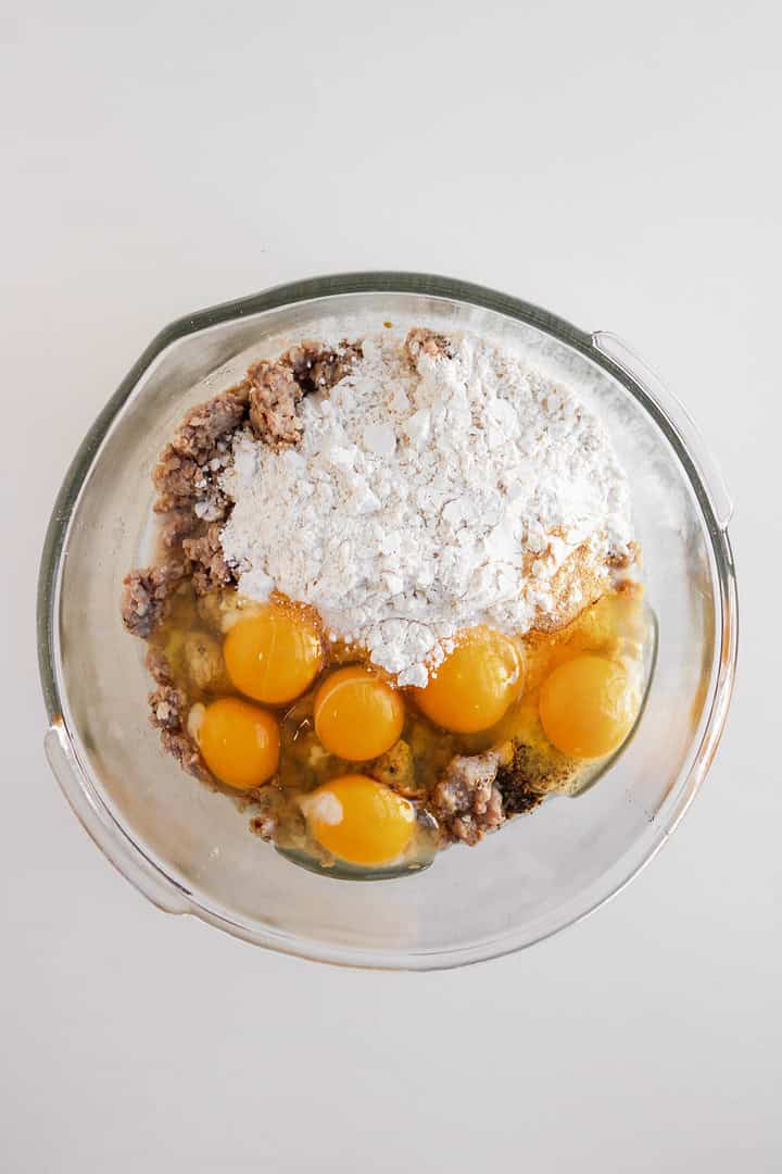 A glass bowl with cracked eggs, flour, and brown mixture on a white surface, ready to be mixed for delicious sausage egg and cheese muffins.