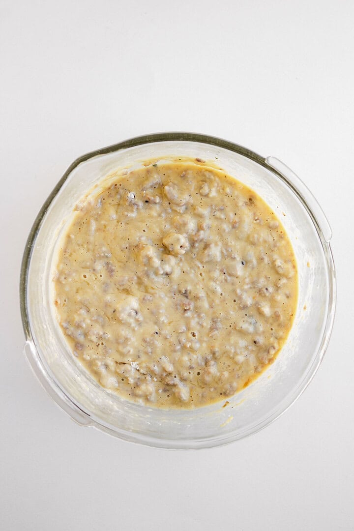 Clear glass bowl filled with a lumpy, yellowish Cheese Breakfast Muffins batter on a white background.