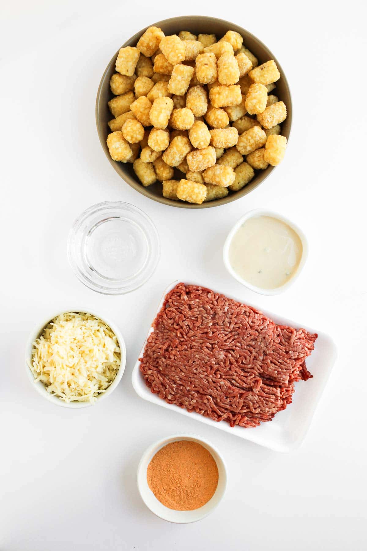 A bowl of Tochos with raw ground beef, shredded cheese, sauce, spices, and a glass of water on a white surface.