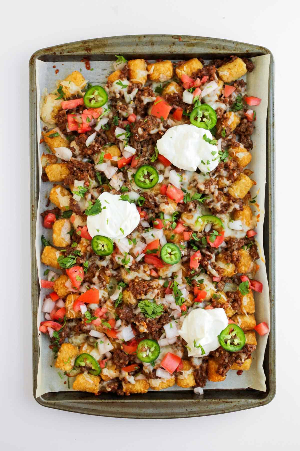 Sheet pan of loaded ground beef tochos topped with cheese, tomatoes, jalape&ntilde;os, onions, and dollops of sour cream.