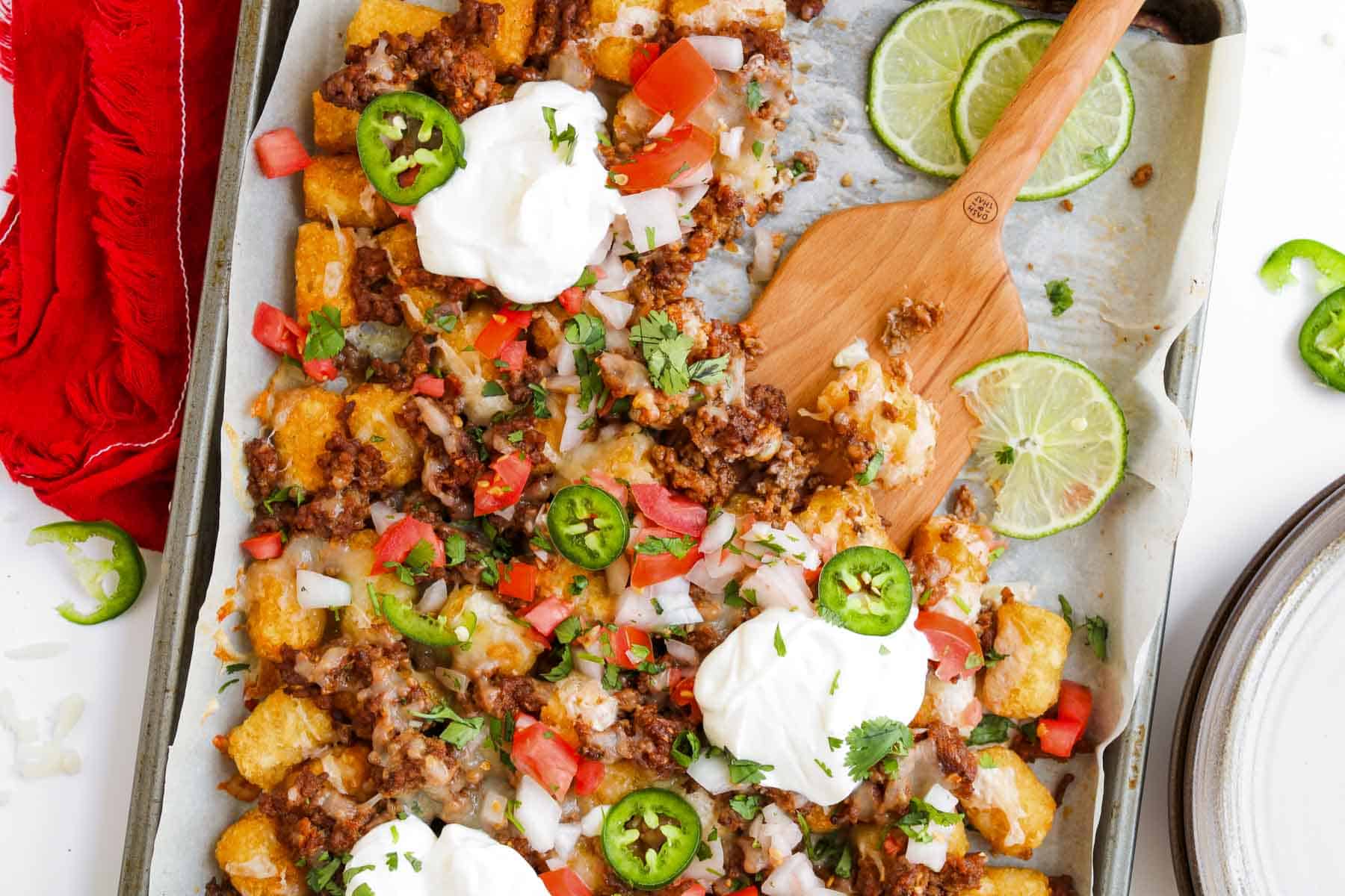 Sheet pan Tochos loaded with crispy tater tots, savory ground beef, tomatoes, jalape&ntilde;os, sour cream, cilantro, and lime slices.