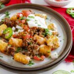 A plate of sheet pan tochos loaded with ground beef, tomatoes, sour cream, and fresh herbs.