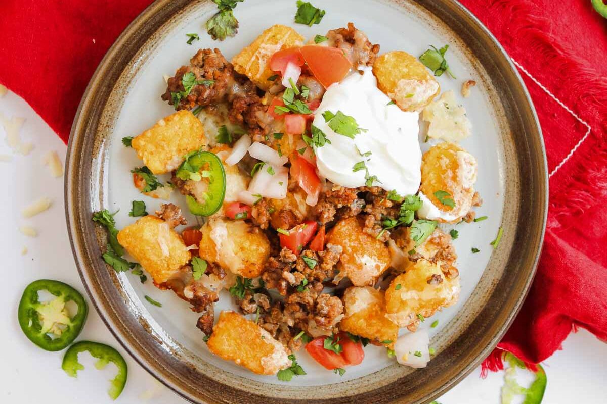 A plate of loaded totchos piled high with ground beef, sour cream, jalape&ntilde;os, tomatoes, and fresh cilantro.