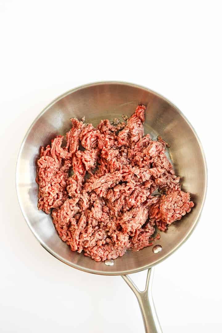 Ground beef in a stainless steel skillet on a white background, ready to be cooked for sheet pan dinners or delicious tochos.
