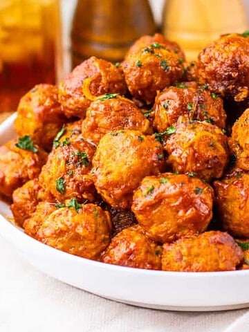 A white bowl filled with glazed meatballs, garnished with herbs, sits on a napkin beside a gold fork.