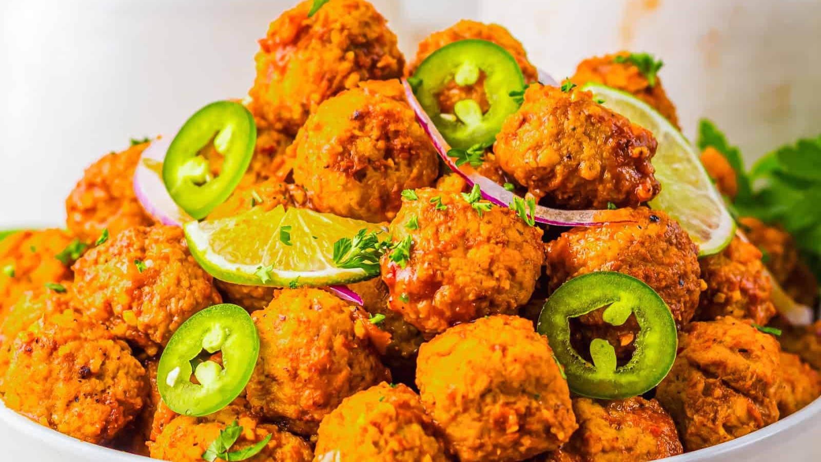 Spicy meatballs garnished with lime slices, jalape&ntilde;o rings, and red onion on a white plate.
