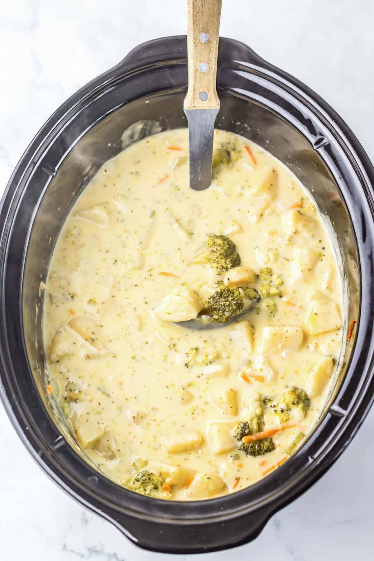 Creamy cheesy broccoli and potato soup simmering in a slow cooker, served with a ladle inside.