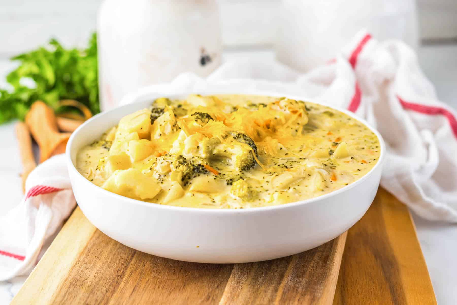 A bowl of creamy slow cooker cheesy broccoli and cheese soup sits on a wooden board with a white towel in the background.