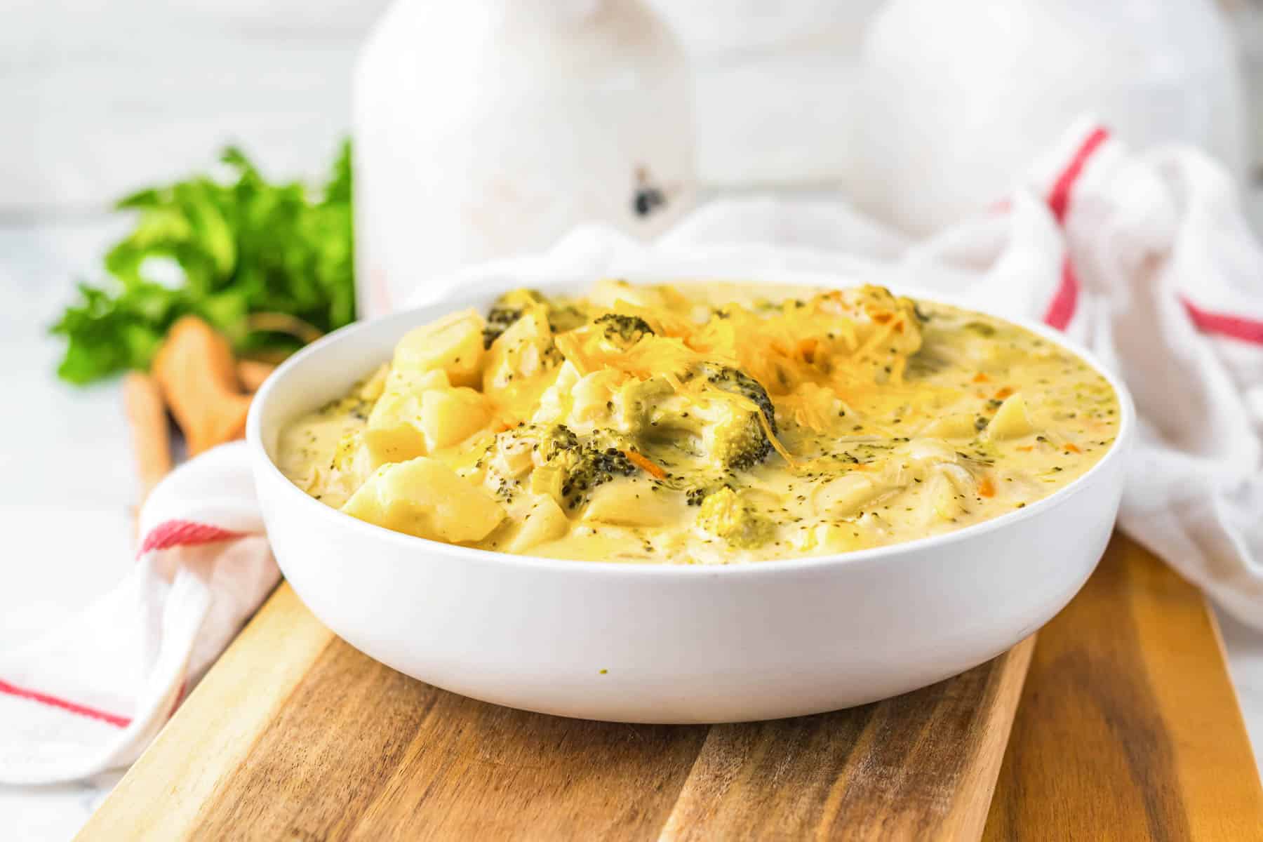 A white bowl of slow cooker cheesy broccoli and cheese soup sits on a wooden board with herbs in the background.