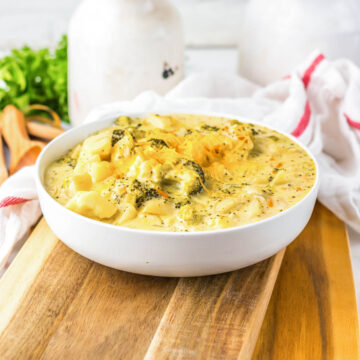 A bowl of creamy cheesy broccoli soup sits on a wooden board, alongside kitchen utensils and a plant in the background—perfect comfort food for slow cooker enthusiasts.