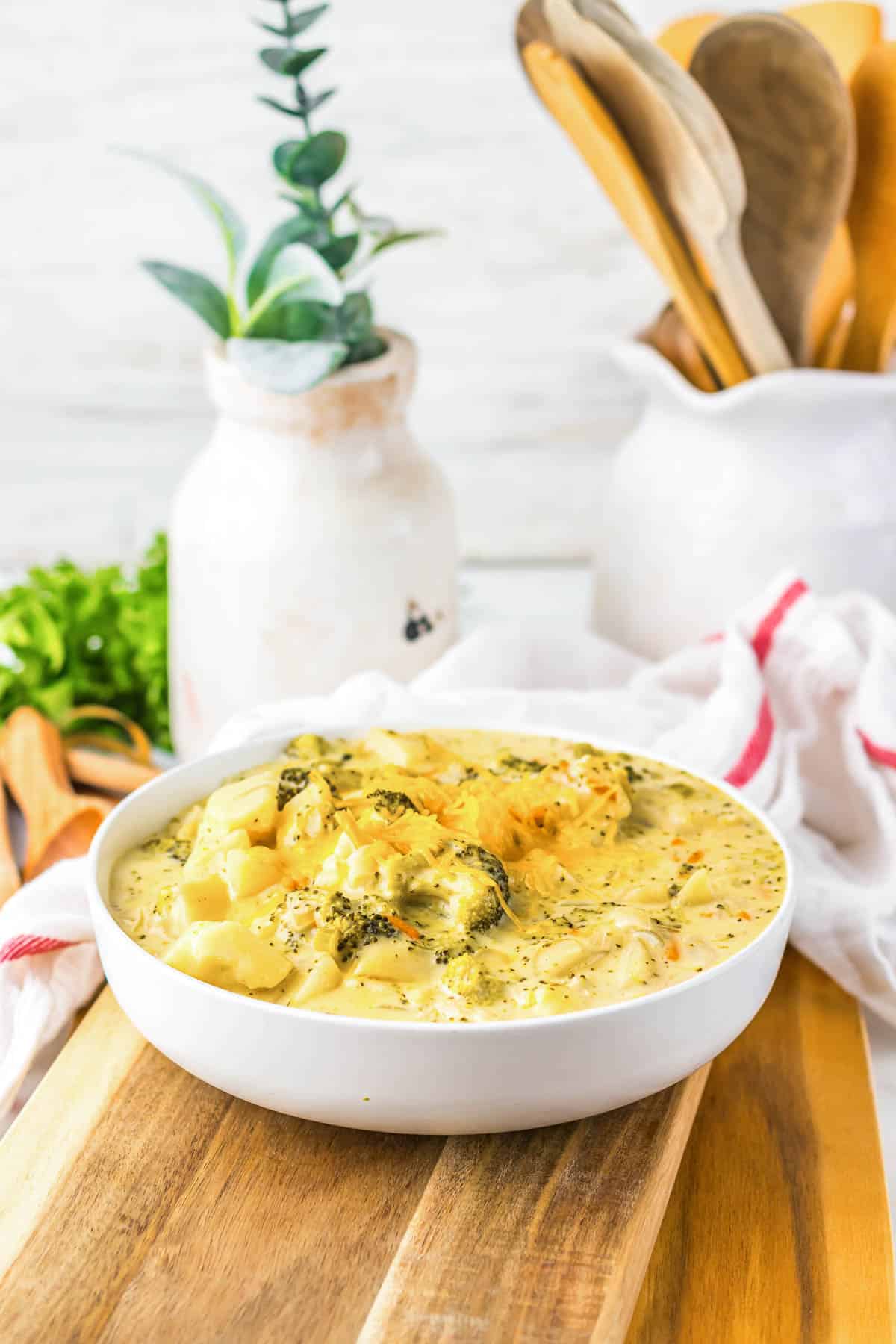 Bowl of creamy broccoli potato soup on a wooden board, with kitchen utensils and decor in the background—a comforting cheesy soup perfect for cozy nights.