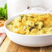 A white bowl filled with creamy potato soup and cheesy broccoli, topped with shredded cheese, sits on a wooden board—perfect for a comforting Slow Cooker meal.
