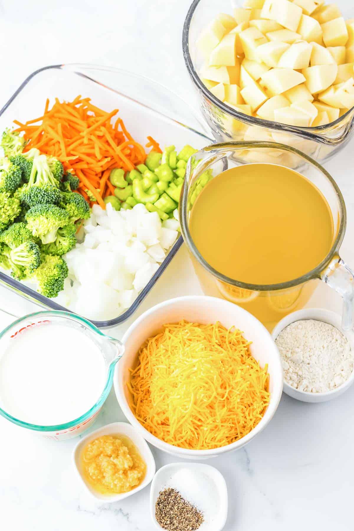 Chopped potatoes, carrots, celery, broccoli, onions, broth, milk, shredded cheese, flour, and seasonings on a counter—everything you need for a hearty slow cooker potato soup with cheesy broccoli.