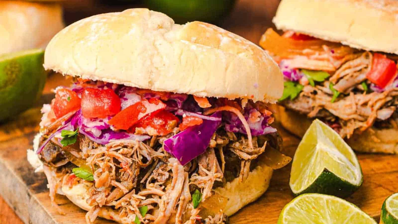 Pulled pork sandwich topped with coleslaw and diced tomatoes, served with lime wedges on a wooden board.