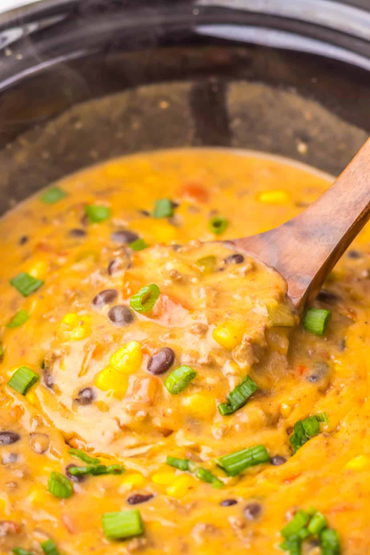Creamy Nacho Soup with corn, black beans, diced tomatoes, and green onions in a pot, stirred with a wooden spoon&mdash;an easy recipe perfect for the slow cooker.