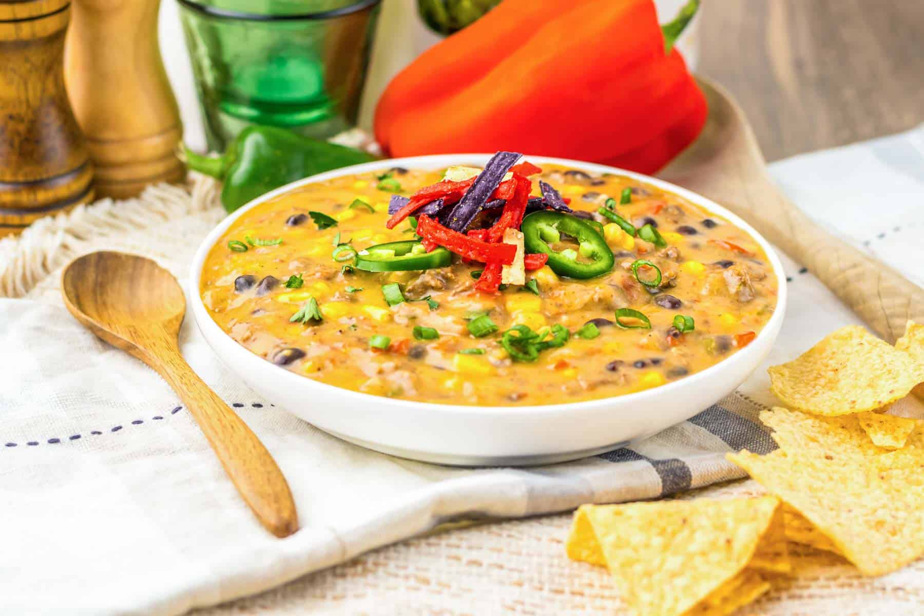 A bowl of cheesy Slow Cooker Nacho Soup with black beans, topped with sliced peppers and tortilla strips, served next to crispy tortilla chips.