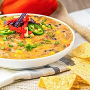 Bowl of cheesy Slow Cooker Nacho Soup with beans, topped with tortilla strips and jalapeños, served alongside tortilla chips and a red bell pepper.