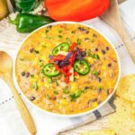 A bowl of cheesy bean dip, inspired by Nacho Soup, topped with jalapeños and tortilla strips, surrounded by chips and fresh vegetables—a perfect recipe for sharing.