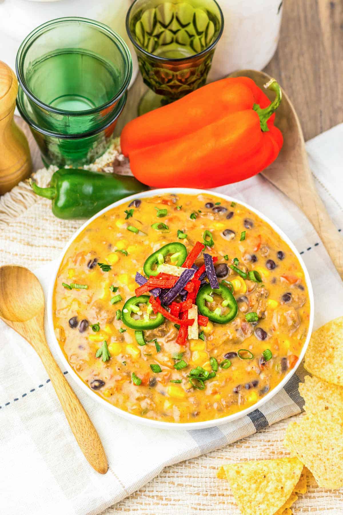 A bowl of cheesy Nacho Soup with black beans, corn, jalape&ntilde;o slices, and tortilla strips, surrounded by fresh peppers&mdash;an irresistible recipe that's perfect for making in your slow cooker.
