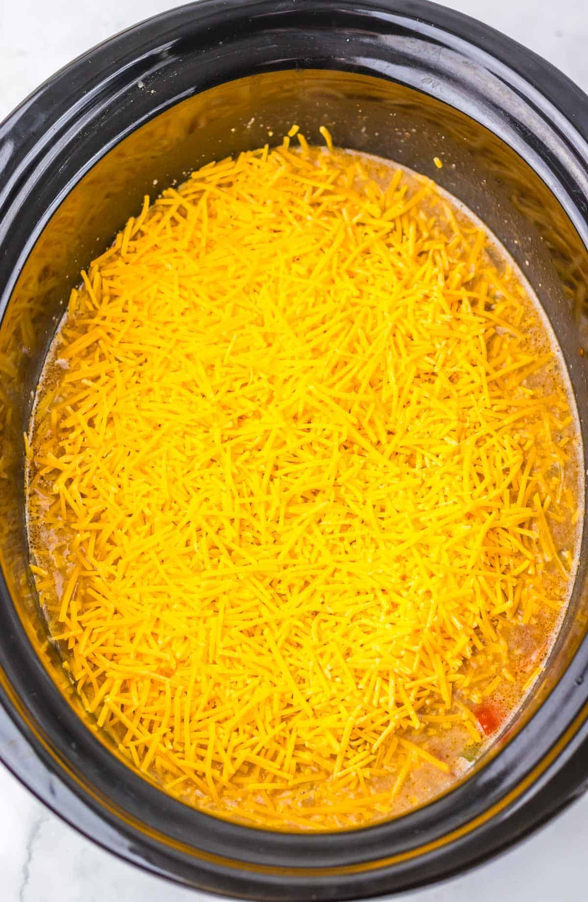 Shredded cheddar cheese tops the ingredients in a slow cooker, ready to be cooked into a delicious nacho soup recipe.