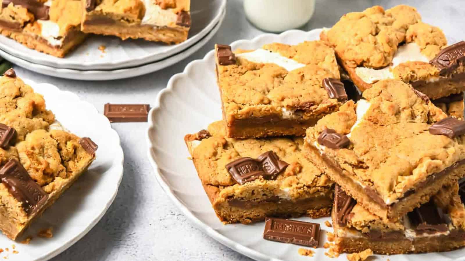 Plates of s&rsquo;mores cookie bars with chocolate pieces and a glass of milk in the background.