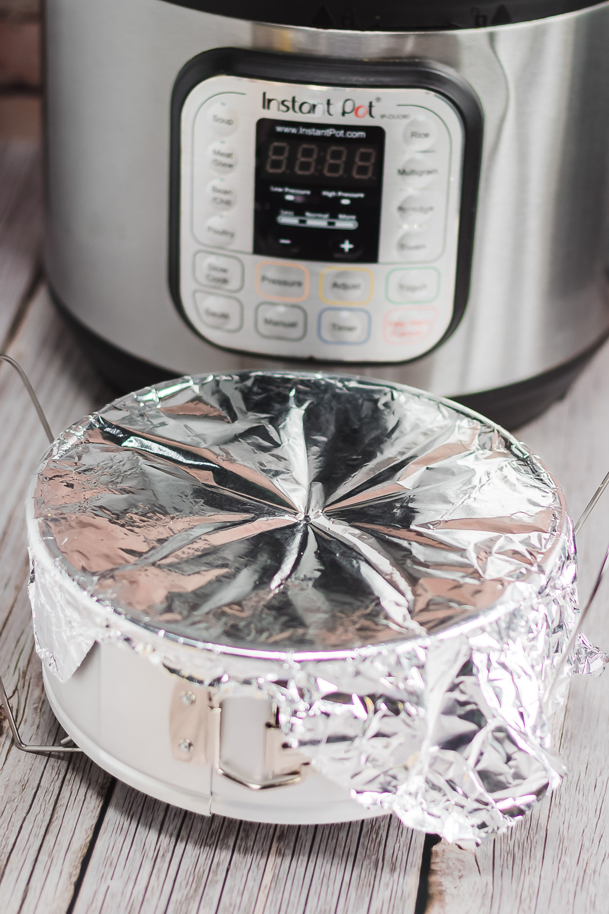 A foil-covered pan sits on a wooden surface in front of an Instant Pot, ready for a delicious Raspberry Lemon Cheesecake recipe.