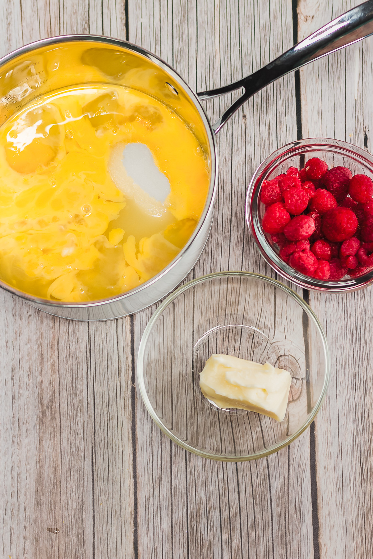 A saucepan with eggs and sugar sits next to bowls of raspberries and butter on a wooden table, perfect for preparing a Raspberry Lemon Cheesecake recipe.