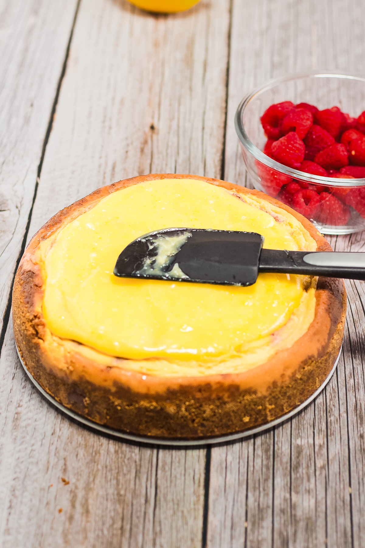 A delicious Raspberry Lemon Cheesecake is topped with yellow lemon curd using a black spatula, next to a bowl of raspberries—a tempting scene for any recipe lover.