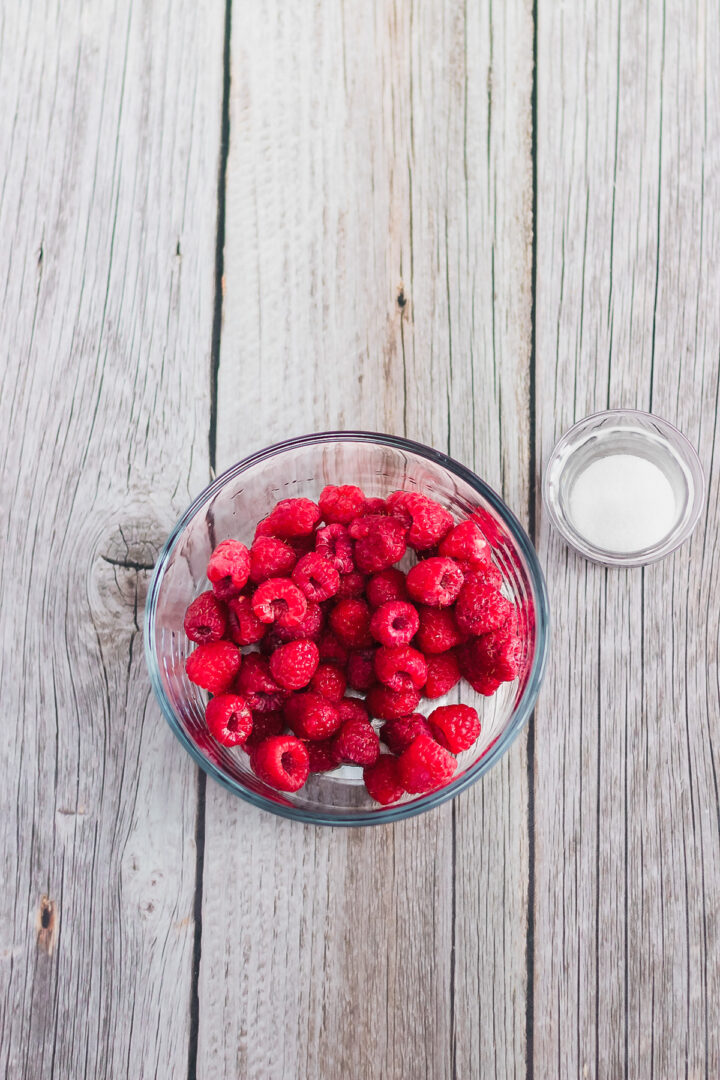 A glass bowl of fresh raspberries and a small dish of sugar on a rustic wooden surface, perfect for topping your favorite Raspberry Lemon Cheesecake recipe.