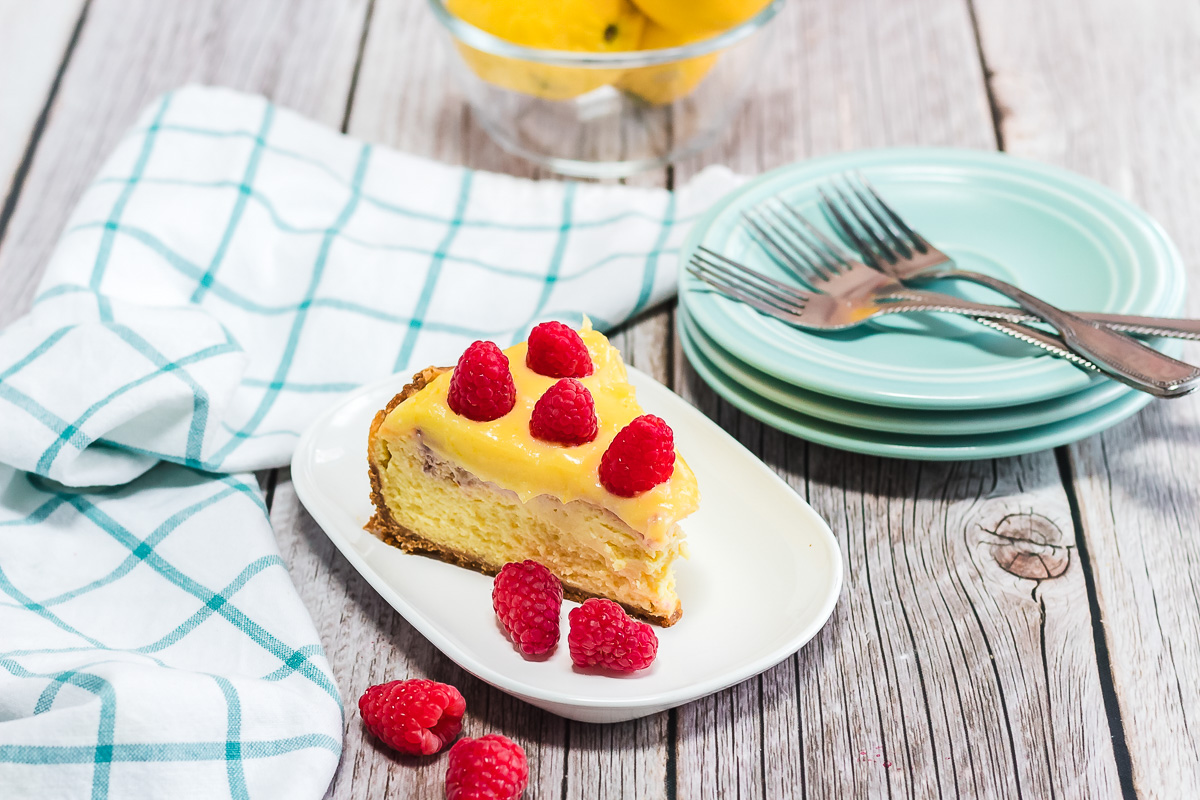 A slice of Raspberry Lemon Cheesecake with raspberries on a plate, next to stacked dishes, forks, and a checkered towel—perfect for trying your favorite Instant Pot recipe.