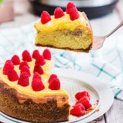 A slice of Raspberry Lemon Cheesecake with yellow topping and raspberries is lifted from the cake; an Instant Pot sits in the background, hinting at the easy recipe behind this delicious dessert.