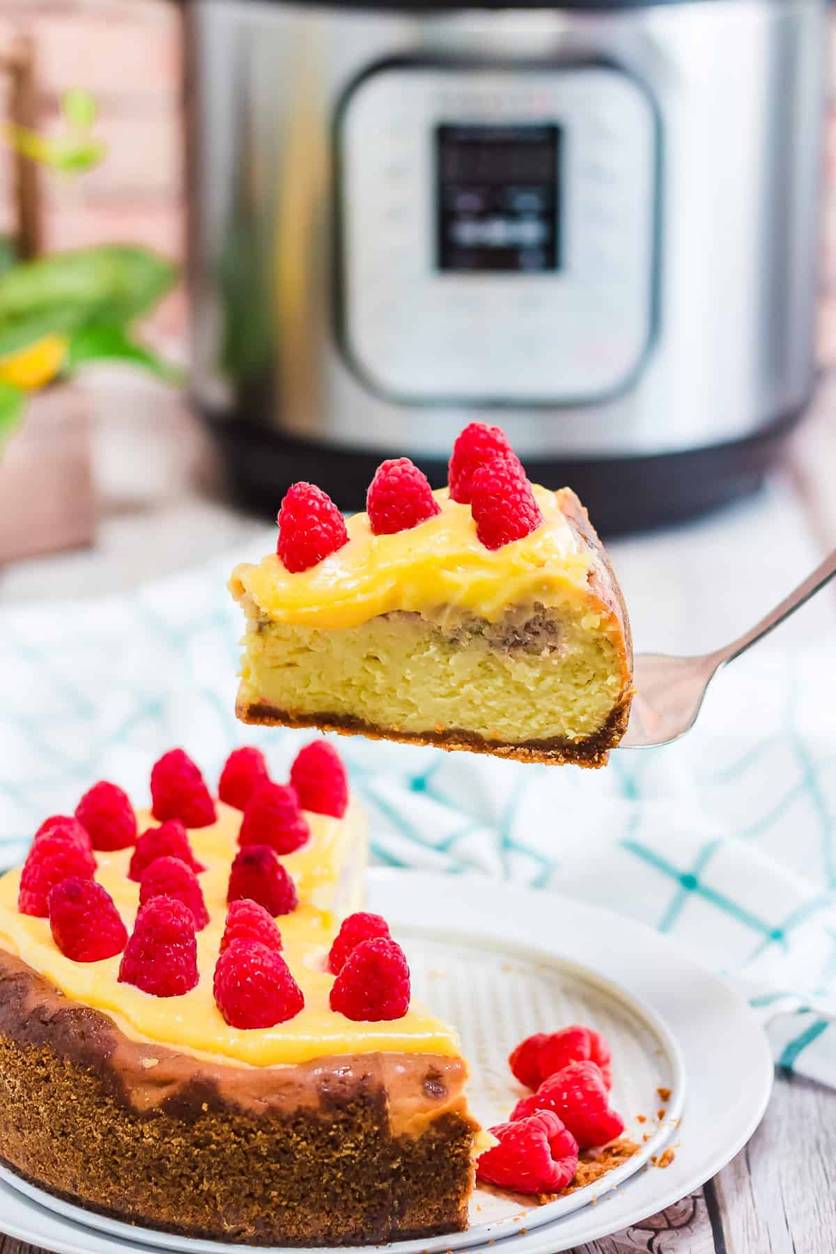 A slice of Raspberry Lemon Cheesecake with yellow topping and raspberries is lifted from the cake; an Instant Pot sits in the background, hinting at the easy recipe behind this delicious dessert.