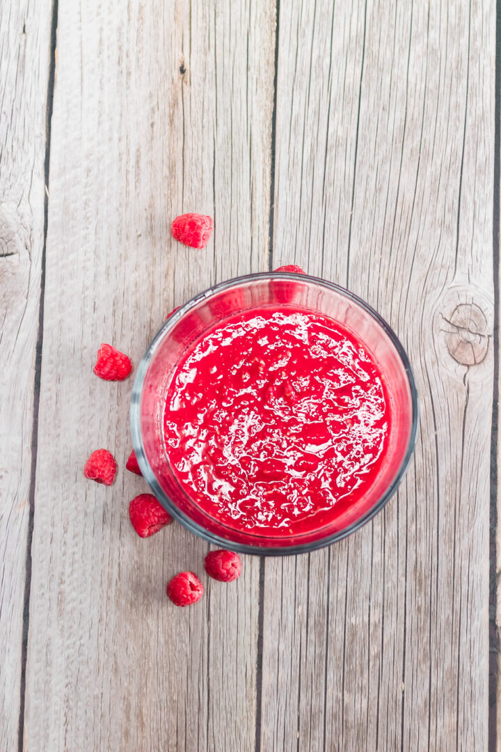 A glass of bright red raspberry smoothie on a wooden surface with scattered raspberries around it, perfect for pairing with a Raspberry Lemon Cheesecake recipe or your next Instant Pot creation.