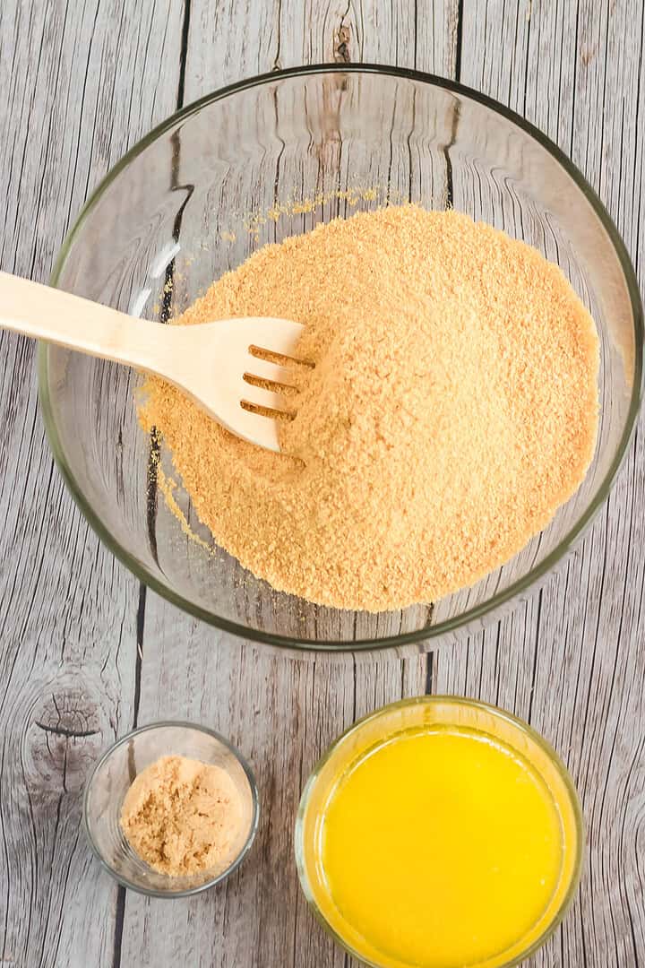 Glass bowl of graham cracker crumbs with a spoon, next to melted butter and brown sugar on a wooden table—the perfect start for your Instant Pot Raspberry Lemon Cheesecake recipe.