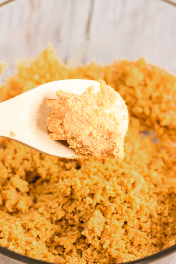 A wooden spoon holds a scoop of crumbly graham cracker mixture above a bowl, ready to become the delicious base for this Raspberry Lemon Cheesecake Instant Pot recipe.