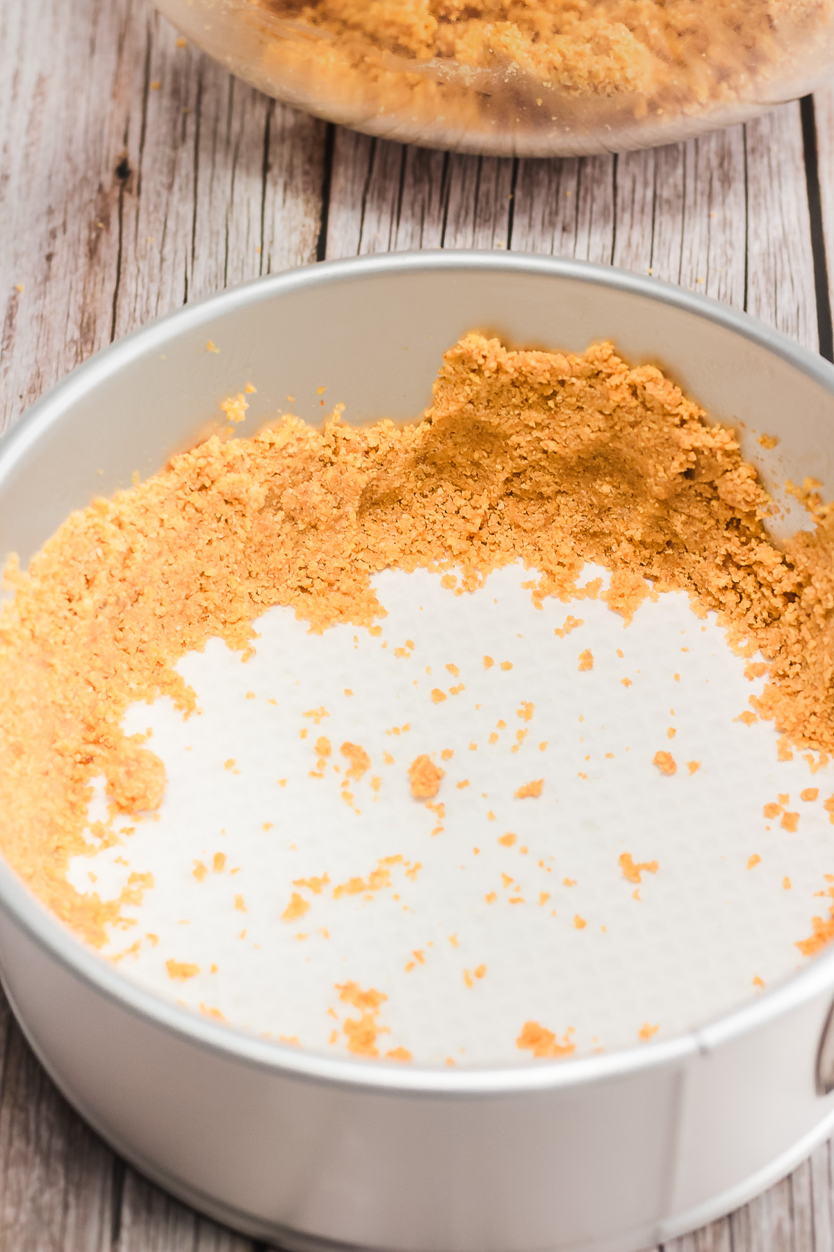 Crushed graham cracker crust pressed into a round cake pan on a wooden surface, ready for a delicious Raspberry Lemon Cheesecake Instant Pot recipe.