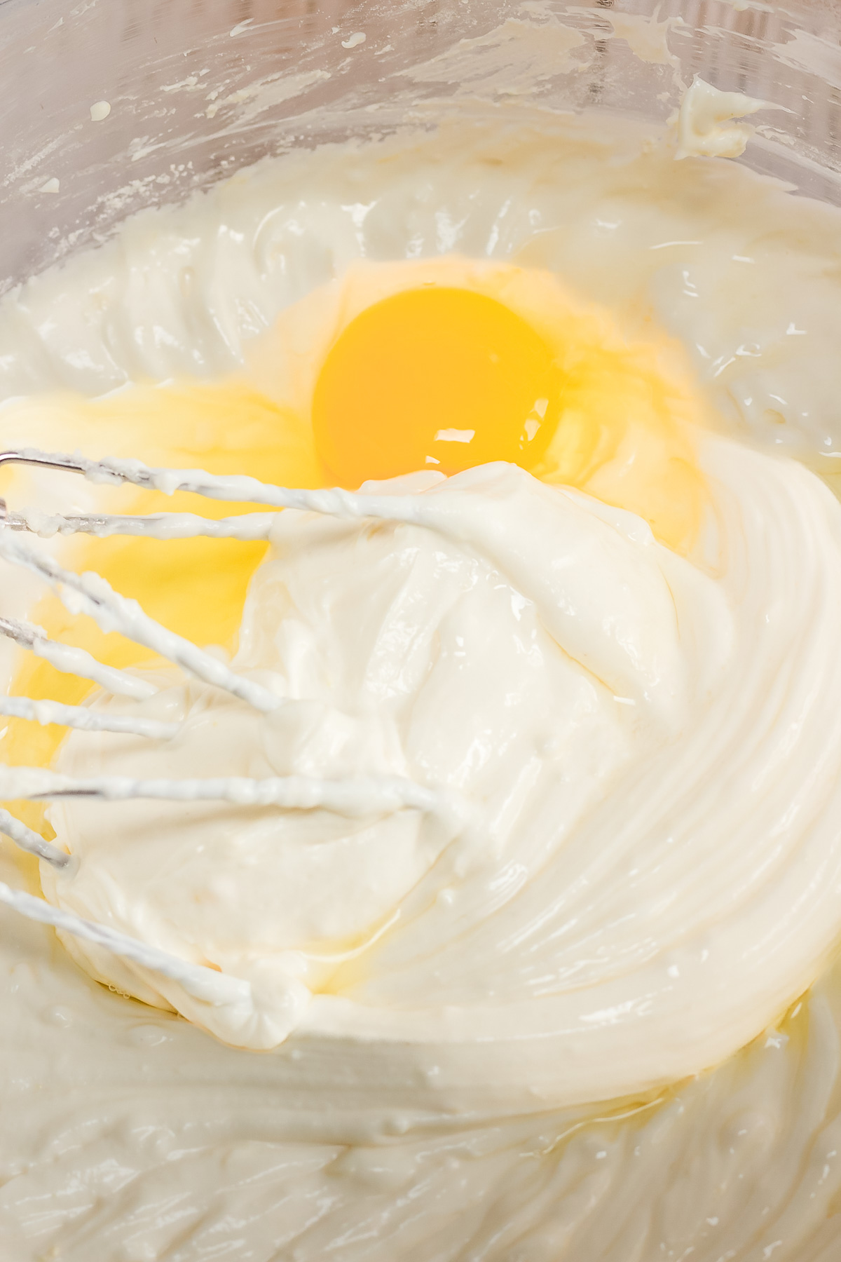 A whisk mixing a raw egg and creamy white batter in a clear bowl, preparing the base for a delicious Raspberry Lemon Cheesecake recipe.
