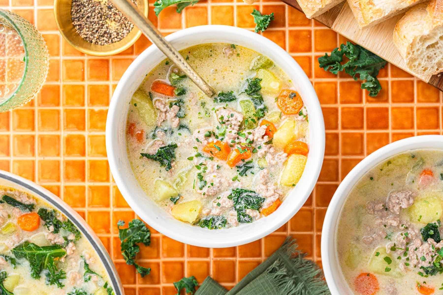 Three bowls of creamy Tuscan Sausage potato soup with ground meat, potatoes, carrots, and kale on an orange tiled surface&mdash;a hearty recipe for any occasion.