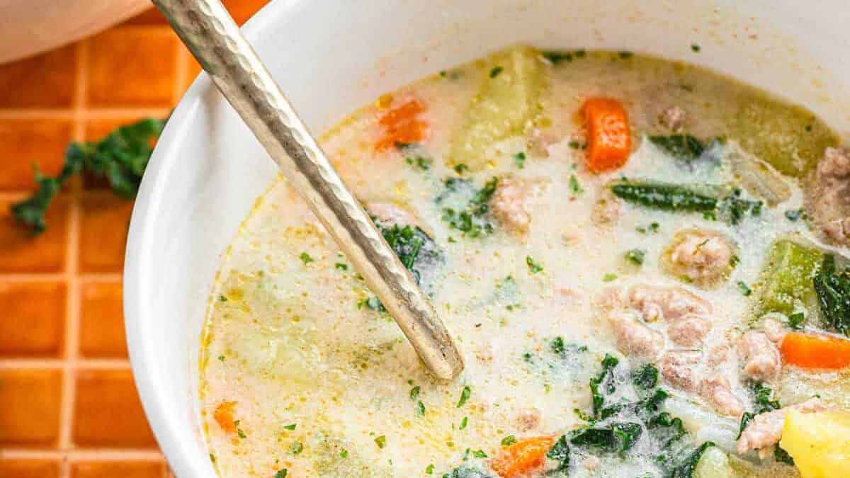 A bowl of creamy Tuscan Sausage and potato soup with ground meat, carrots, kale, and a spoon inside&mdash;an irresistible recipe perfect for cozy nights.