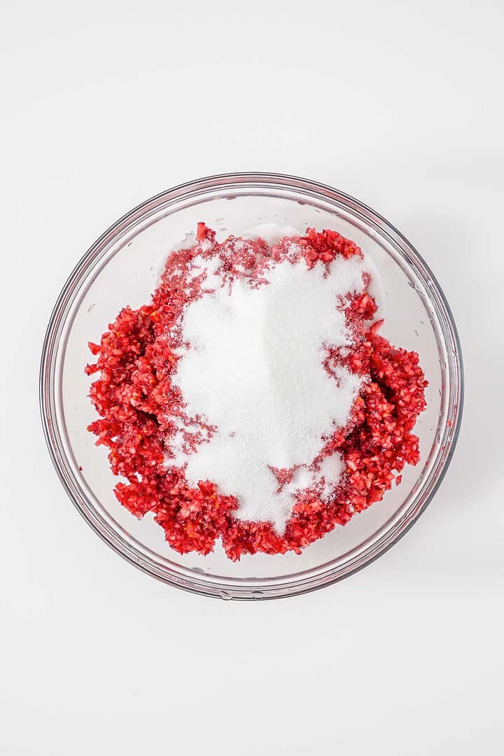 A glass bowl with chopped red fruit and a mound of white sugar on top, set on a white surface.