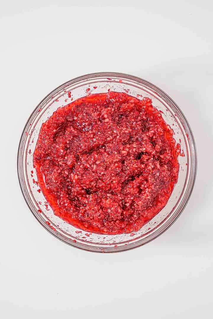 A glass bowl filled with a chunky red mixture, resembling cranberry relish, on a white surface.